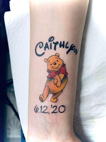 Colour Tattoo von Winnie Puuh mit dem Namen und dem Datum auf dem Unterarm, liebevolles Erinnerungstattoo in warmen Farben, gestochen bei Talisman Tattoo in Raeren, Ostbelgien, nahe Aachen, Eupen und Luxemburg