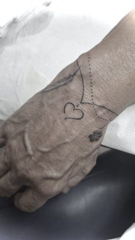 Fineline Handgelenk Tattoo mit Herz, Feder und Dotwork Linie, minimalistisches Linework Tattoo am Handgelenk und Handrücken, feines Symbol Tattoo in Black, gestochen bei Talisman Tattoo – Sabine Jacobs, exklusives Tattoo Studio in Belgien nahe Aachen, NRW, Eupen, Sankt Vith, Luxemburg, Europa, internationale Tattoo Kundschaft