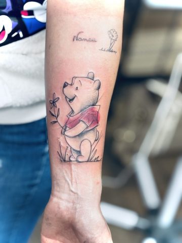 Zartes Winnie Puuh Tattoo im Aquarell- und Sketch-Stil auf dem Unterarm, Comic-Motiv mit rotem Shirt, Blume und feiner Linienführung, liebevolles Erinnerungs-Tattoo gestochen bei Talisman Tattoo in Raeren, Ostbelgien, nahe Aachen, Eupen und Luxemburg