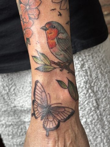 Neotraditional Colour Tattoo eines Rotkehlchens auf einem Ast mit Blättern und Schmetterling am Unterarm, warme Farben und feine Schattierungen, gestochen bei Talisman Tattoo in Raeren, Belgien, nahe Aachen, Eupen und Luxemburg