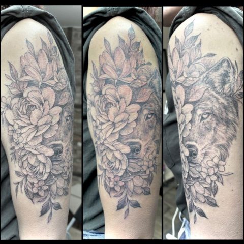 Fineline Wolf Tattoo in Black & Grey mit floralen Elementen und detailreichen Blumen auf dem Oberarm einer Frau.