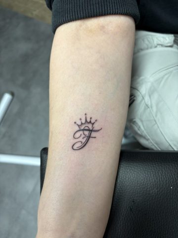 Filigranes Initialen Tattoo mit Krone auf dem Unterarm im Fine Line und Lettering Stil.